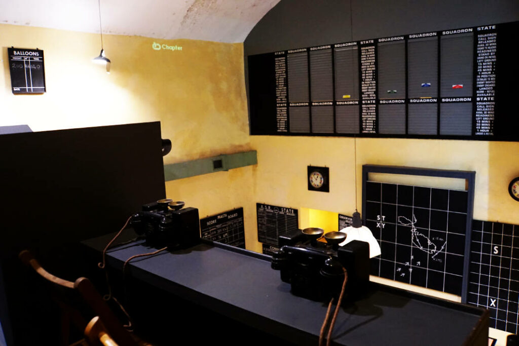 Ein historischer Blick in die Einsatzzentrale des Lascaris War Room in Malta mit alten Telefonen auf einem Tresen und großen Informationstafeln an den gelben Wänden, die Flugzeugbewegungen und Einsatzbereitschaften anzeigen.