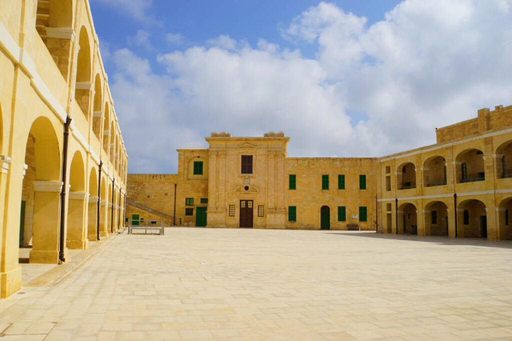 Der weitläufige, gepflasterte Innenhof des Fort St. Elmo in Valletta, Malta, ist von historischen, sandgelben Steingebäuden mit markanten Arkadenbögen und grünen Fensterläden unter einem leicht bewölkten blauen Himmel umgeben.
