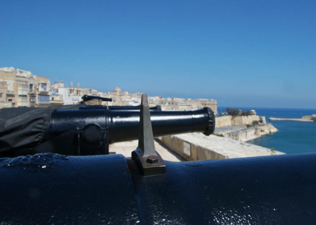 Nahaufnahme einer schwarzen Kanone der Saluting Battery in Valletta, Malta, mit Blick über weitere Kanonenrohre hinweg auf den Hafen und die historische Stadtkulisse unter blauem Himmel.