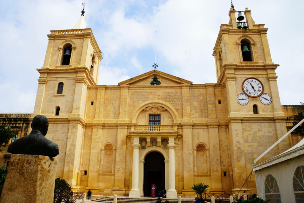 Die St. John’s Co-Cathedral in Valletta, Malta, präsentiert sich mit ihrer sandfarbenen Barockfassade, zwei markanten Glockentürmen mit Uhren und einem zentralen Eingangsportal unter einem bewölkten Himmel. Im Vordergrund links ist die Rückansicht einer dunklen Büste auf einem Steinsockel zu sehen.