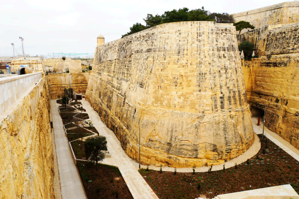 Die massiven, sandsteinfarbenen Stadtmauern von Valletta in Malta ragen steil über einem tiefen Graben auf, der heute mit geometrisch angelegten Gehwegen und kleinen Grünanlagen gestaltet ist.