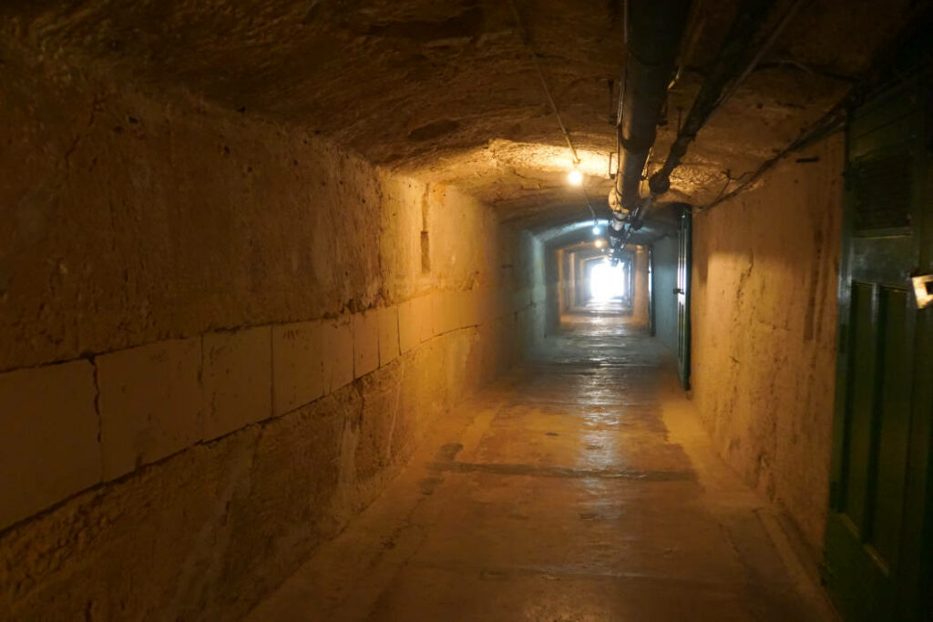 Ein schmaler, schwach beleuchteter unterirdischer Tunnel mit rauen Steinwänden und einer Rohrleitung an der Decke führt zum Lascaris War Room in Valletta, Malta. Am Ende des langen Ganges ist ein heller Lichtschein zu sehen.