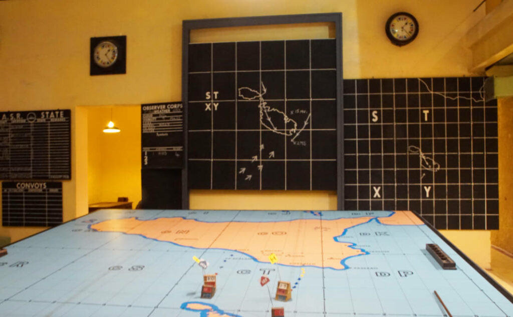 Ein historischer War Room in Valletta, Malta, zeigt einen großen Kartentisch mit Markierungen für Truppenbewegungen sowie Wandtafeln mit Koordinatennetzen und strategischen Diagrammen.