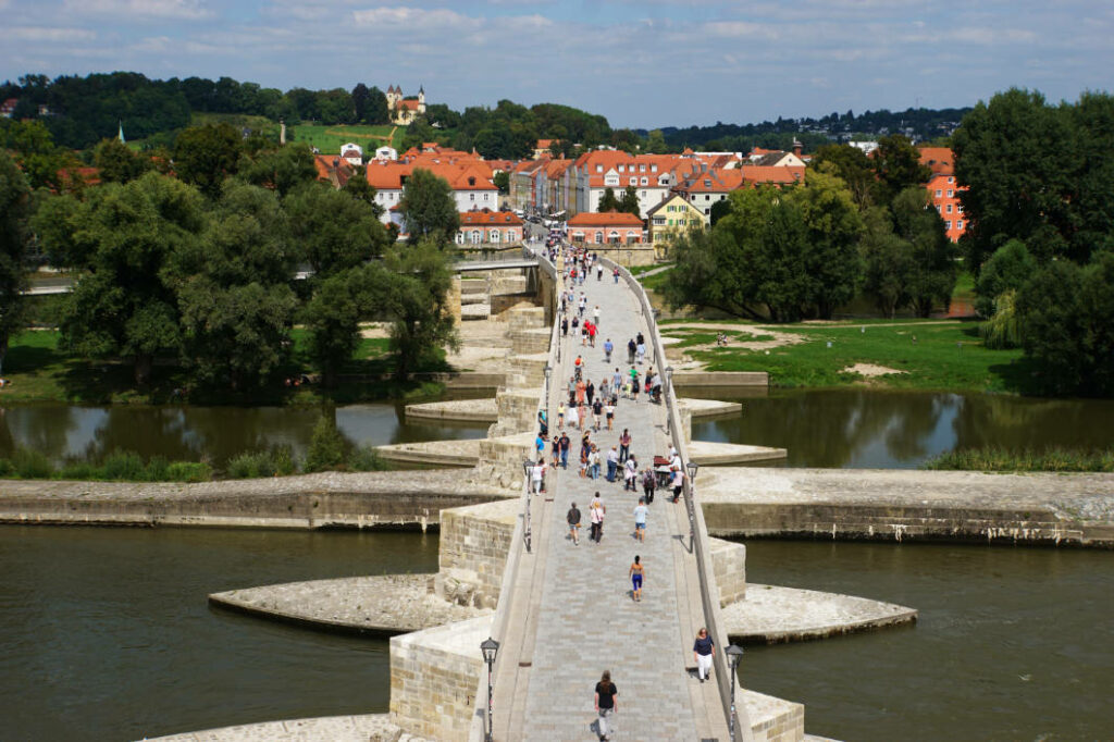 Blick von oben auf die Steinerne Brücke in Regensburg, auf der zahlreiche Menschen in Richtung des Stadtteils Stadtamhof mit seinen charakteristischen Häusern mit roten Dächern spazieren.