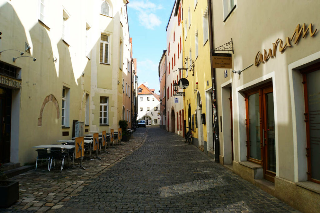 Eine schmale, gepflasterte Gasse in der Regensburger Altstadt, gesäumt von mehrstöckigen, hellgelben und weißen Häusern mit kleinen Außengastronomie-Tischen auf der linken Seite.