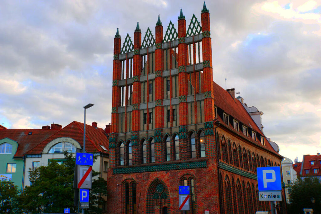 Das Alte Rathaus in Stettin präsentiert sich als markanter Backsteinbau der Backsteingotik mit einer reich verzierten Schaufassade aus roten Ziegeln und grünen Akzenten unter einem bewölkten Himmel. Das historische Gebäude am „Roten Weg“ ist von modernen Straßenschildern und benachbarten Wohnhäusern mit roten Ziegeldächern umgeben.