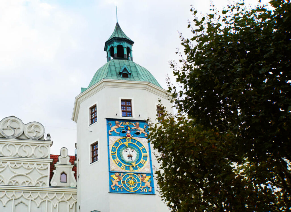 Die astronomische Uhr am Turm des Herzogsschlosses in Stettin zeigt ein blaues Zifferblatt mit goldenen Details und einer zentralen Sonne, eingebettet in die weiße Fassade unter einer grünen Kuppel. Das historische Bauwerk ist eine Station auf dem „Roten Weg“ durch die Stadt.