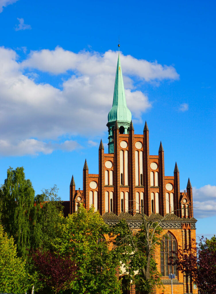 Die Backsteinfassade der Kirche St. Peter und Paul in Stettin ragt mit ihrem markanten Stufengiebel und der grünen Turmspitze vor einem blau-weißen Wolkenhimmel empor.