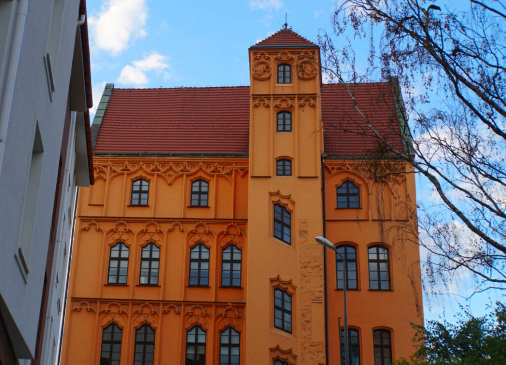 Die Außenansicht des historischen Loitzenhofs in Stettin zeigt ein mehrstöckiges, orangefarbenes Gebäude mit spätgotischen Maßwerkverzierungen und einem markanten Treppenturm unter einem roten Ziegeldach.