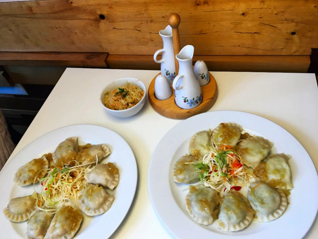 Zwei weiße Teller mit Stettiner Piroggen, garniert mit Krautsalat und Zwiebeln, stehen neben einer kleinen Schüssel Salat und einem Menagen-Set auf einem weißen Tisch vor einer Holzwand.