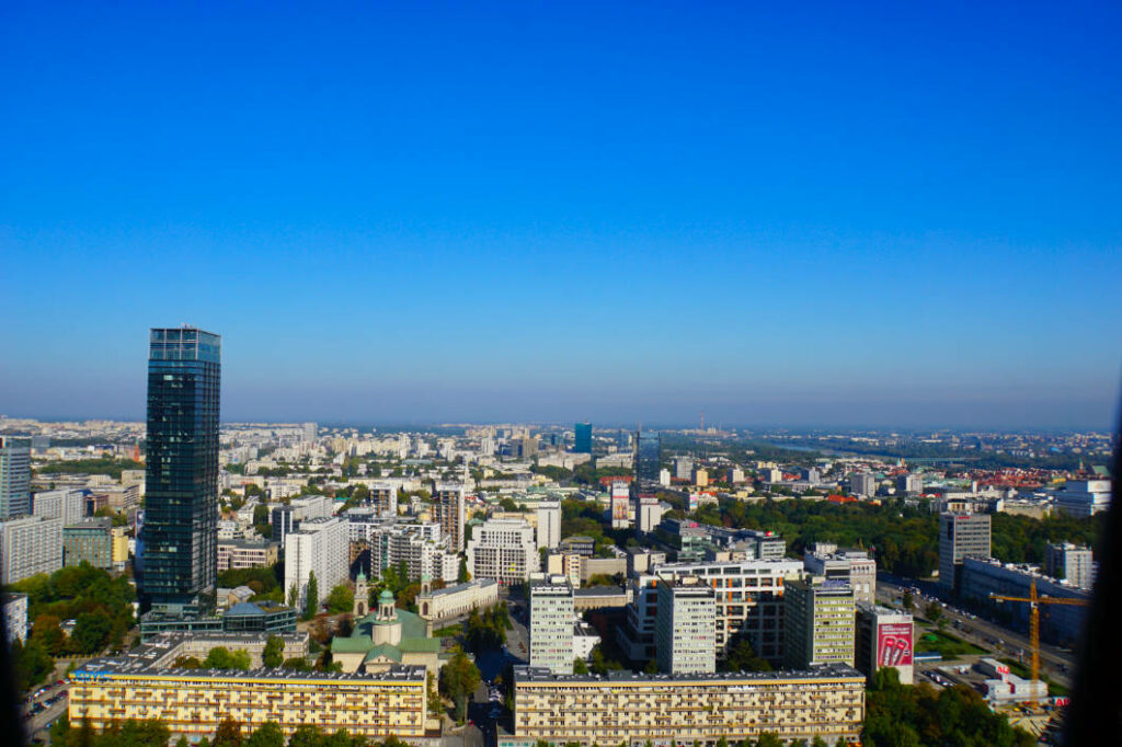 Weitläufiger Blick vom Kultur- und Wissenschaftspalast über das herbstliche Warschau mit dem markanten dunklen Wolkenkratzer Cosmopolitan Twarda 4 im Vordergrund unter strahlend blauem Himmel.
