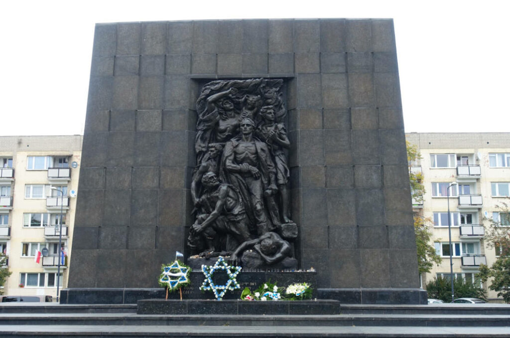 Das Denkmal der Helden des Ghettos in Warschau zeigt eine monumentale, dunkle Steinwand mit einer zentralen Bronzeskulptur kämpfender Figuren, vor der Gedenkkränze in Form von Davidsternen niedergelegt wurden. Das Mahnmal befindet sich auf dem Gelände des POLIN Museums der Geschichte der polnischen Juden vor einer Kulisse aus modernen Wohngebäuden.