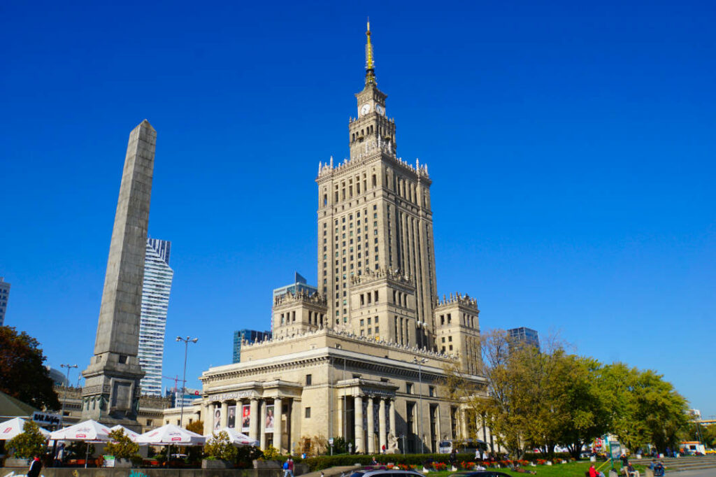 Der monumentale Kultur- und Wissenschaftspalast in Warschau ragt unter strahlend blauem Himmel hinter einem steinernen Obelisken und herbstlichen Bäumen empor.