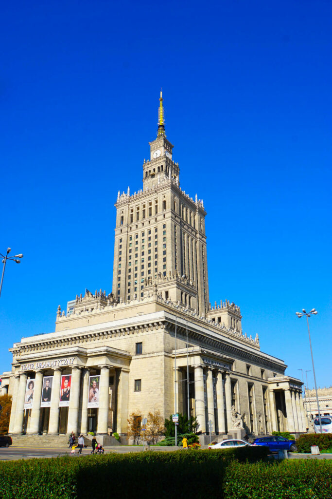 Der monumentale Kultur- und Wissenschaftspalast in Warschau ragt unter einem strahlend blauen Himmel empor, wobei die markante Architektur im stalinistischen Stil und der hohe Turm mit seiner goldenen Spitze detailliert zur Geltung kommen. Im Vordergrund sind der Eingangsbereich des Teatr Dramatyczny mit Säulen und Plakaten sowie eine gepflegte Grünanlage zu sehen.