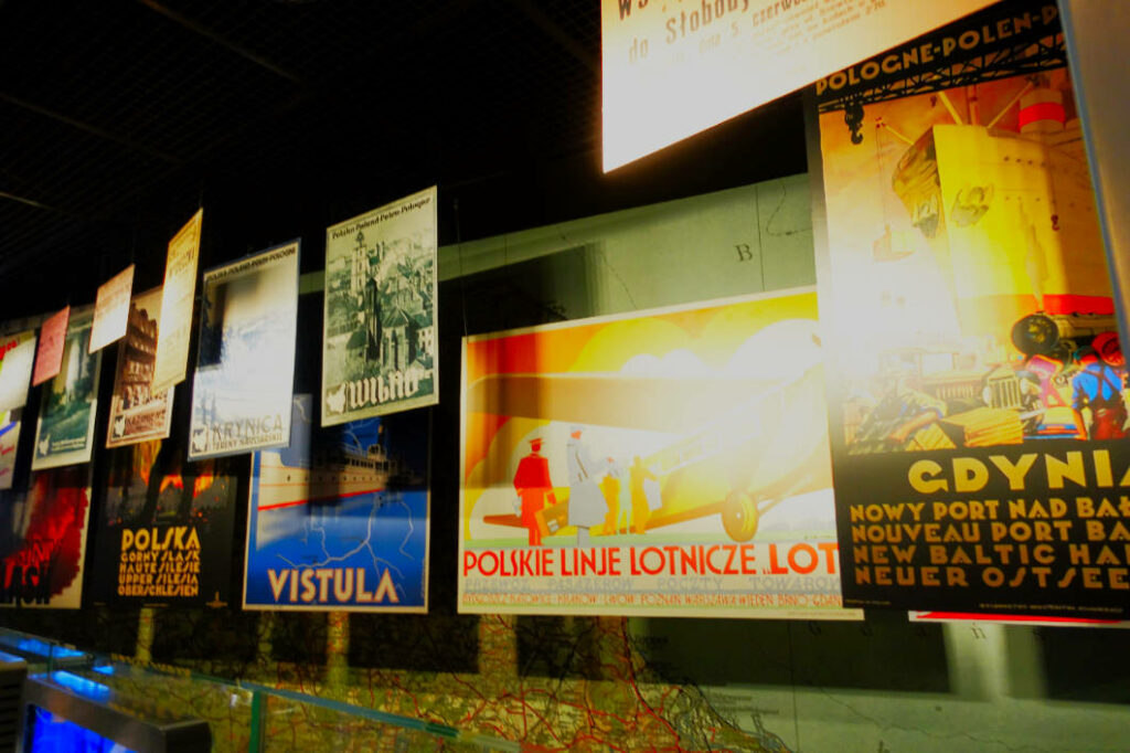 Eine Ausstellung im POLIN Museum der Geschichte der polnischen Juden in Warschau zeigt historische Reiseplakate für polnische Städte und Regionen, darunter Gdynia, Wilno und die Weichsel. Die farbenfrohen Grafiken hängen in Reihen vor einer dunklen Decke und beleuchten verschiedene Reiseziele der jüdischen Bevölkerung im Vorkriegspolen.