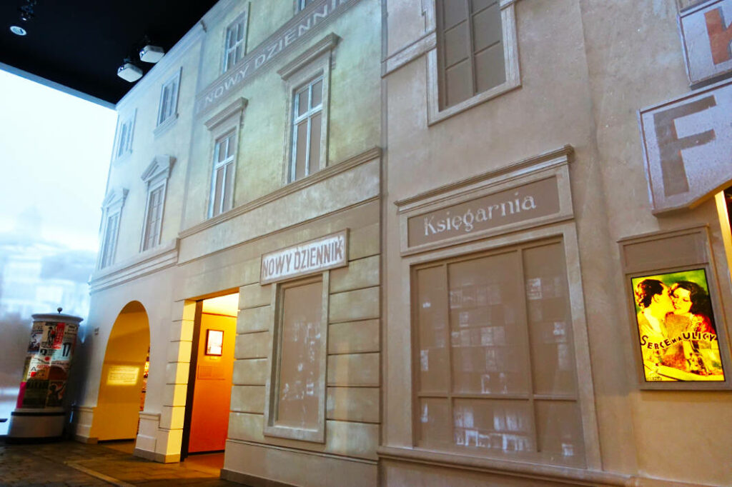 Eine museale Rekonstruktion eines historischen Straßenzugs im POLIN Museum der Geschichte der polnischen Juden in Warschau zeigt Fassaden mit polnischen Beschriftungen wie „Księgarnia“ (Buchhandlung) und „Nowy Dziennik“. Die Ausstellung vermittelt durch Kulissen von Geschäften und Wohnhäusern sowie ein beleuchtetes Filmplakat einen Eindruck des jüdischen Alltagslebens im Vorkriegspolen.