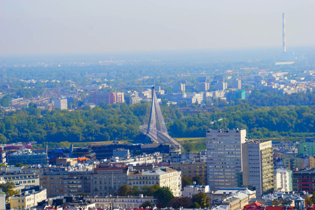 Blick vom Kultur- und Wissenschaftspalast über die Stadt Warschau auf die markante Świętokrzyski-Brücke, die über die Weichsel führt. Im Hintergrund erstreckt sich das Stadtbild mit zahlreichen Wohngebäuden, Grünflächen und einem hohen, rot-weiß gestreiften Schornstein am Horizont.