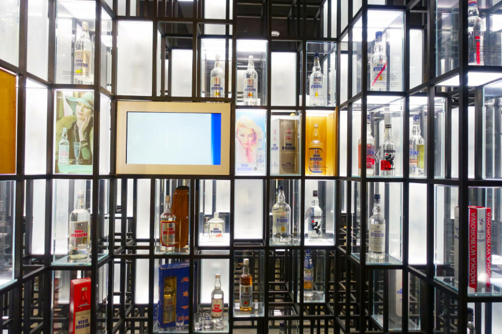 Eine moderne, beleuchtete Wanderausstellung im Polnischen Wodka Museum in Warschau zeigt zahlreiche historische und aktuelle Wodkaflaschen in einem geometrischen, gitterartigen Regalsystem mit integrierten Bildschirmen.