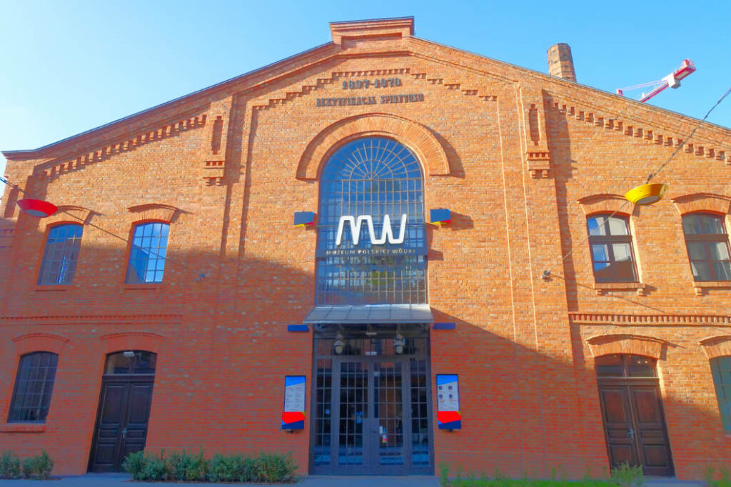 Die Außenansicht des Polnischen Wodka Museums in Warschau zeigt ein historisches Backsteingebäude mit einem großen Rundbogenfenster, über dem das moderne weiße Logo „MW“ prangt. Das Gebäude ist Teil der ehemaligen Koneser-Wodkafabrik und zeichnet sich durch seine charakteristische rote Ziegelarchitektur mit symmetrisch angeordneten Fenstern und Türen aus.