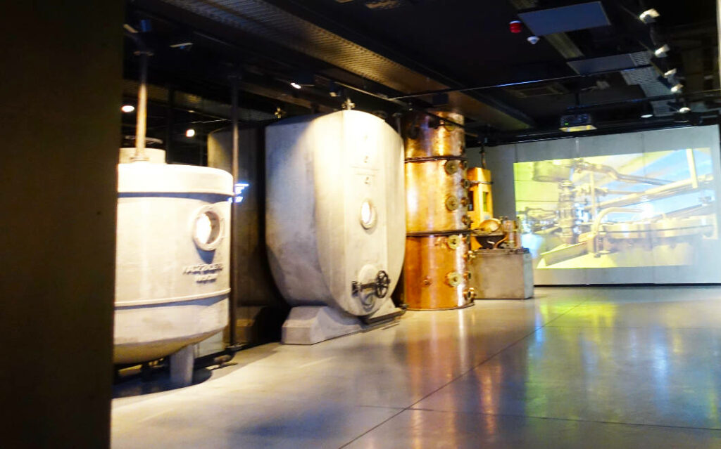 Ausstellungsraum im Polnischen Wodka Museum in Warschau mit historischen Brennereigeräten wie großen weißen Tanks und einer kupfernen Destillationskolonne vor einer Videoprojektion.