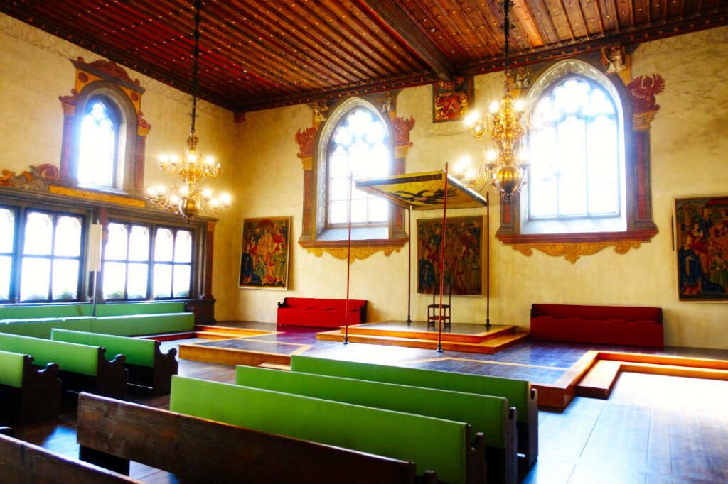 Der historische Reichssaal im Alten Rathaus von Regensburg mit hölzerner Decke, prunkvollen Kronleuchtern, Wandteppichen und einem Thronsessel unter einem Baldachin für den Kaiser des Immerwährenden Reichstags. Im Vordergrund stehen grüne und braune Sitzbänke für die Gesandten vor einer erhöhten hölzernen Plattform.