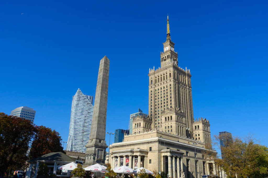 Der Warschauer Kulturpalast ragt unter strahlend blauem Himmel neben einem hohen Steinobelisken und modernen Wolkenkratzern wie dem Złota 44 empor.