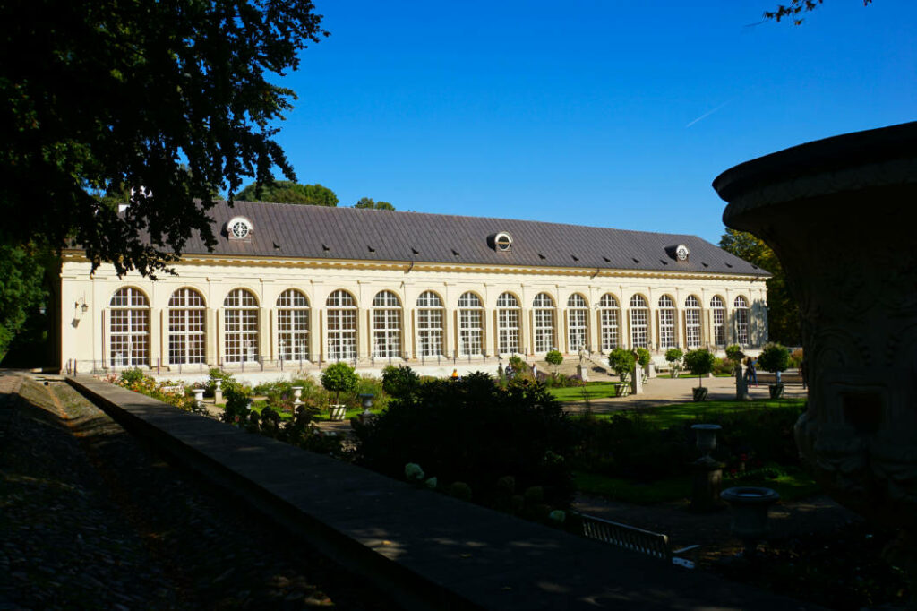 Die Alte Orangerie im Warschauer Łazienki-Park präsentiert sich an einem sonnigen Sommertag mit ihrer langen, hellen Fassade und den charakteristischen hohen Rundbogenfenstern hinter einer gepflegten Gartenanlage.
