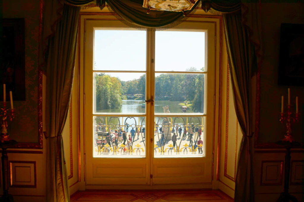 Blick aus einem prunkvollen Fenster eines Palastes in Warschau an einem sonnigen Sommertag auf einen See und zahlreiche Spaziergänger im Park. Das Fenster ist von schweren grünen Vorhängen und goldenen Verzierungen eingerahmt, während draußen eine grüne Parklandschaft mit Wasser zu sehen ist.