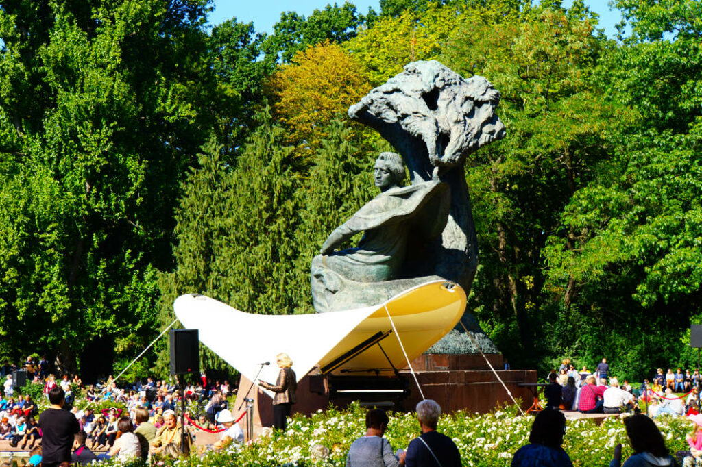 Ein Publikum versammelt sich an einem sonnigen Sonntag im Sommer im Warschauer Łazienki-Park zu einem Klavierkonzert vor dem berühmten Chopin-Denkmal, das den Komponisten unter einer stilisierten Weide zeigt. Ein weißes Sonnensegel überspannt den Flügel und die Pianistin auf der Bühne vor der monumentalen Bronzestatue.