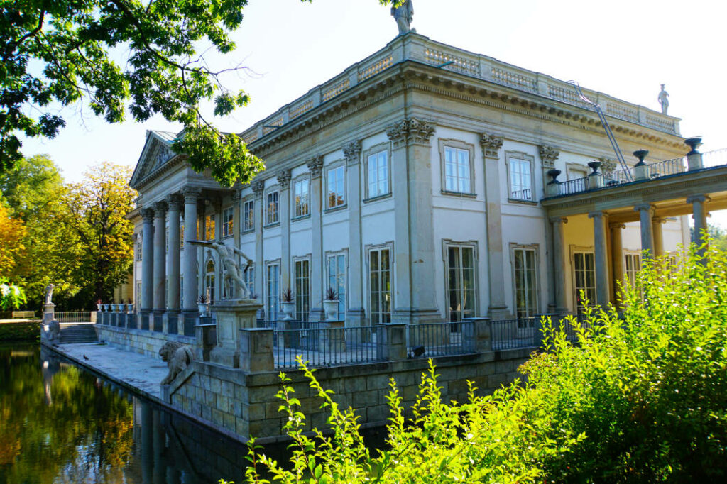 Der klassizistische Łazienki-Palast in Warschau spiegelt sich an einem sonnigen Sommertag im ruhigen Wasser des umliegenden Sees, flankiert von grünen Bäumen und antiken Skulpturen.
