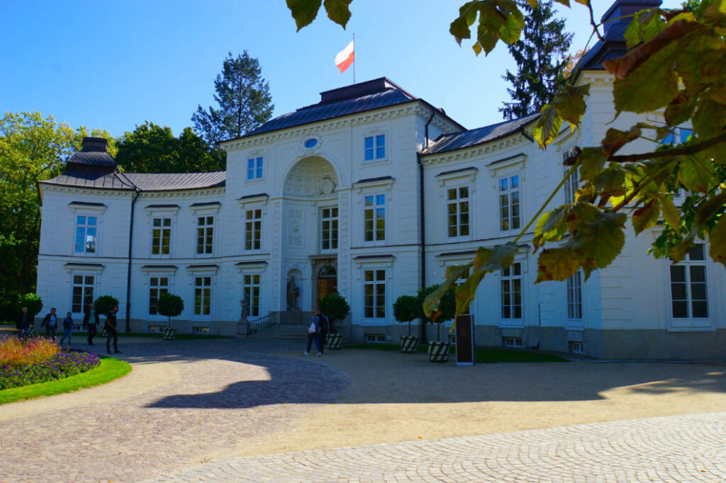 Der weiße, klassizistische Myślewicki-Palast in Warschau unter strahlend blauem Himmel an einem sonnigen Sommertag, umgeben von grünen Bäumen und einer gepflasterten Auffahrt mit Besuchern.