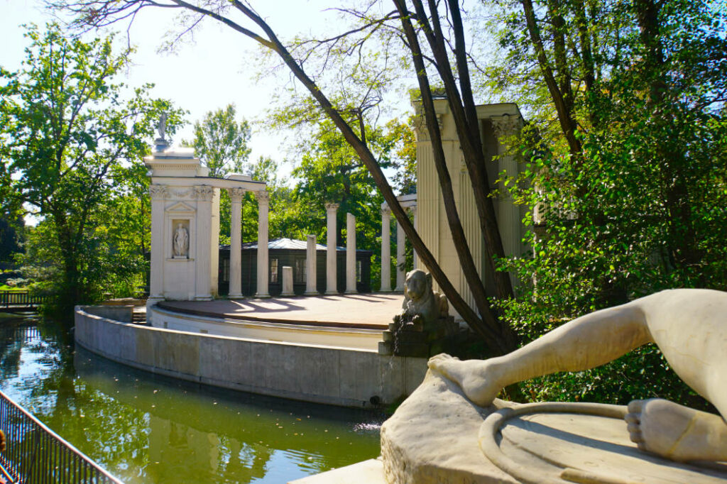 Das antike Amphitheater im Warschauer Łazienki-Park an einem sonnigen Sommertag, mit einer von Säulen gesäumten Bühne am Wasser und einer Steinstatue im Vordergrund.