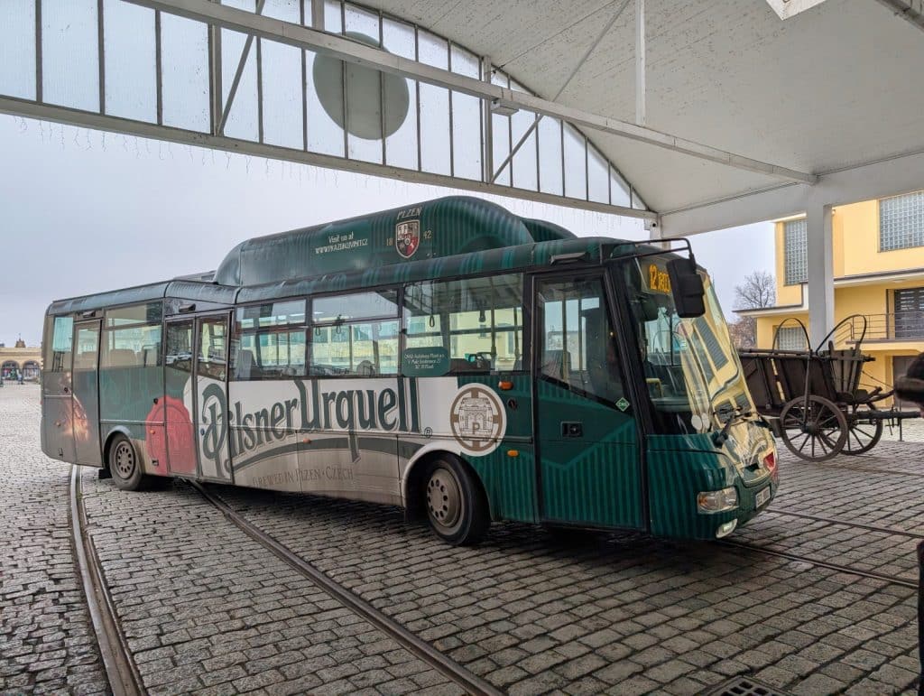 Ein grün-weißer Pilsner Urquell Bus für Brauereiführungen steht auf Kopfsteinpflaster unter einer überdachten Station der Pilsen Brauerei, neben einem historischen Pferdewagen.