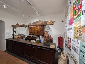 Eine historische Ladeneinrichtung im Ethnografischen Museum der Region Pilsen mit einem dunklen Holztresen, Apothekerschränken und zwei großen, dekorativen Holzfischen, die über der Theke hängen. An der rechten Wand sind zahlreiche bunte, historische Werbeplakate angebracht, während auf dem Tresen alte Waagen, Gläser und Gefäße stehen.