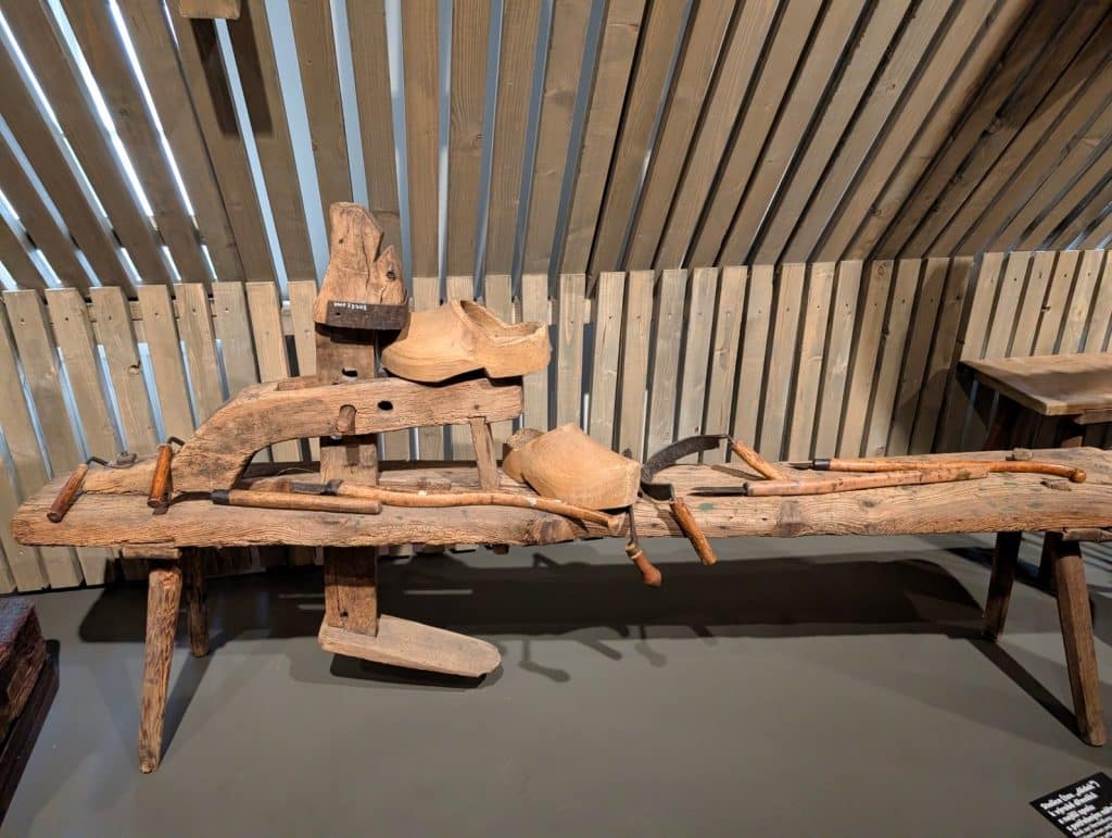 Ein historischer Arbeitsplatz zur traditionellen Herstellung von Holzschuhen im Ethnografischen Museum der Region Pilsen, bestehend aus einer hölzernen Schnitzbank, verschiedenen handgefertigten Werkzeugen und zwei unfertigen Holzschuhen.
