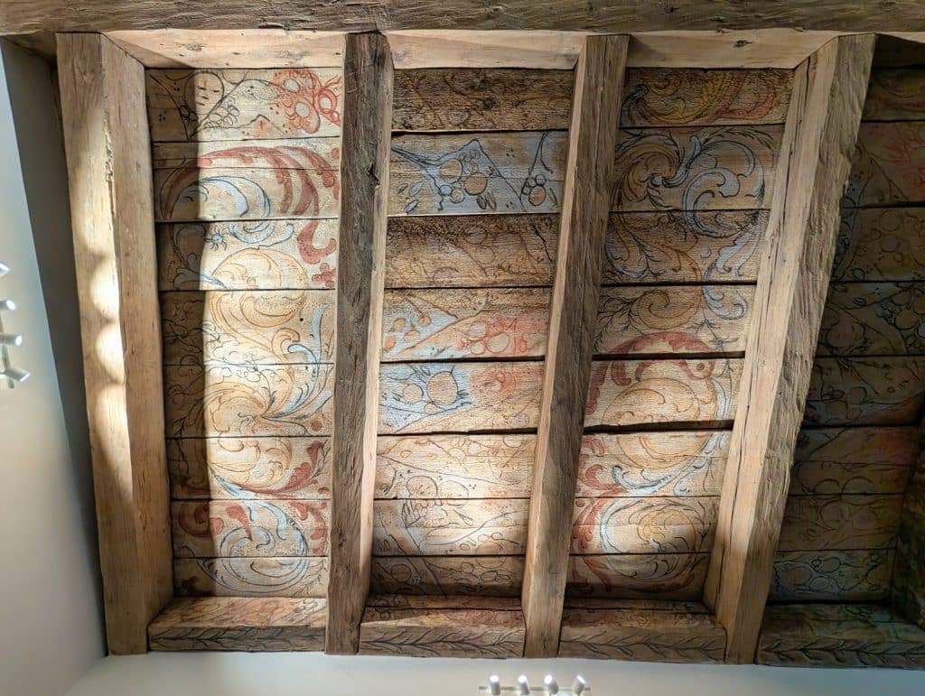 Eine historische Holzdecke im Ethnografischen Museum der Region Pilsen zeigt kunstvoll bemalte Paneele mit floralen Ornamenten und Rankenmotiven zwischen massiven, rustikalen Holzbalken.
