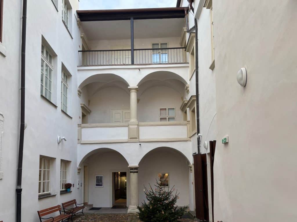 Der helle Innenhof des Ethnografischen Museums der Region Pilsen zeigt eine mehrstöckige Arkadenfassade mit Rundbögen und Säulen auf dem Kopfsteinpflaster. An der linken weißen Hauswand stehen zwei Holzbänke unter Sprossenfenstern.