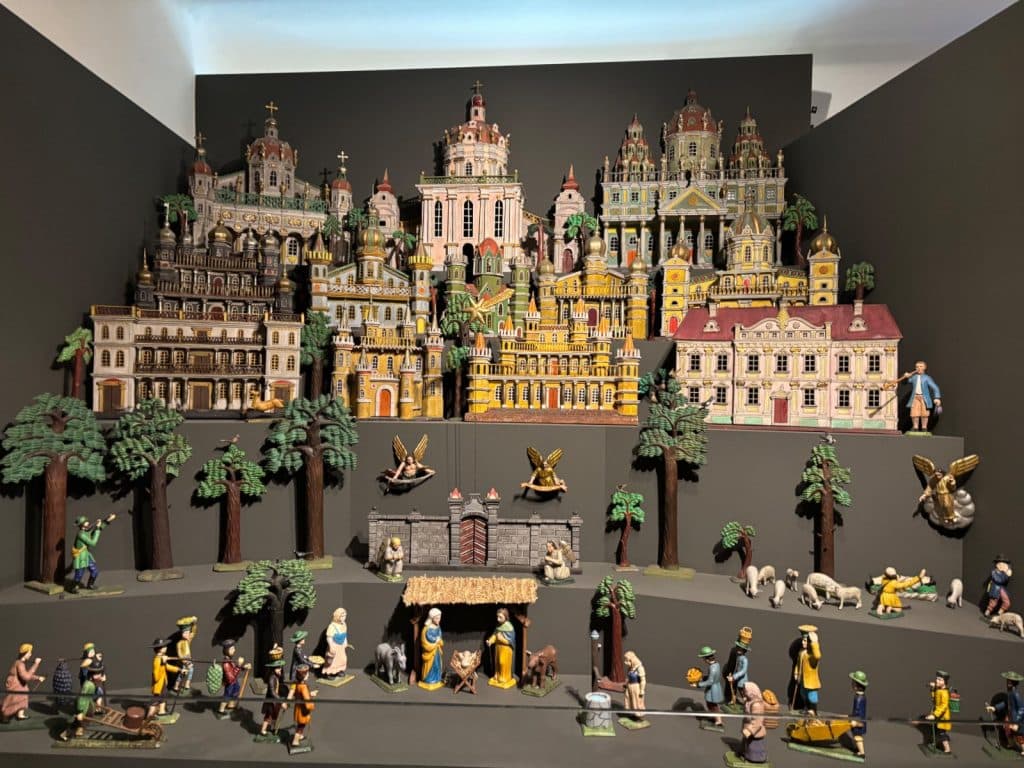 Eine detailreiche, mehrstöckige Weihnachtskrippe im Ethnografischen Museum der Region Pilsen zeigt im Vordergrund die heilige Familie im Stall, umgeben von zahlreichen Figuren und Tieren, während im Hintergrund eine prächtige, fantasievolle Stadtlandschaft mit bunten Kirchen und Palästen aufragt.