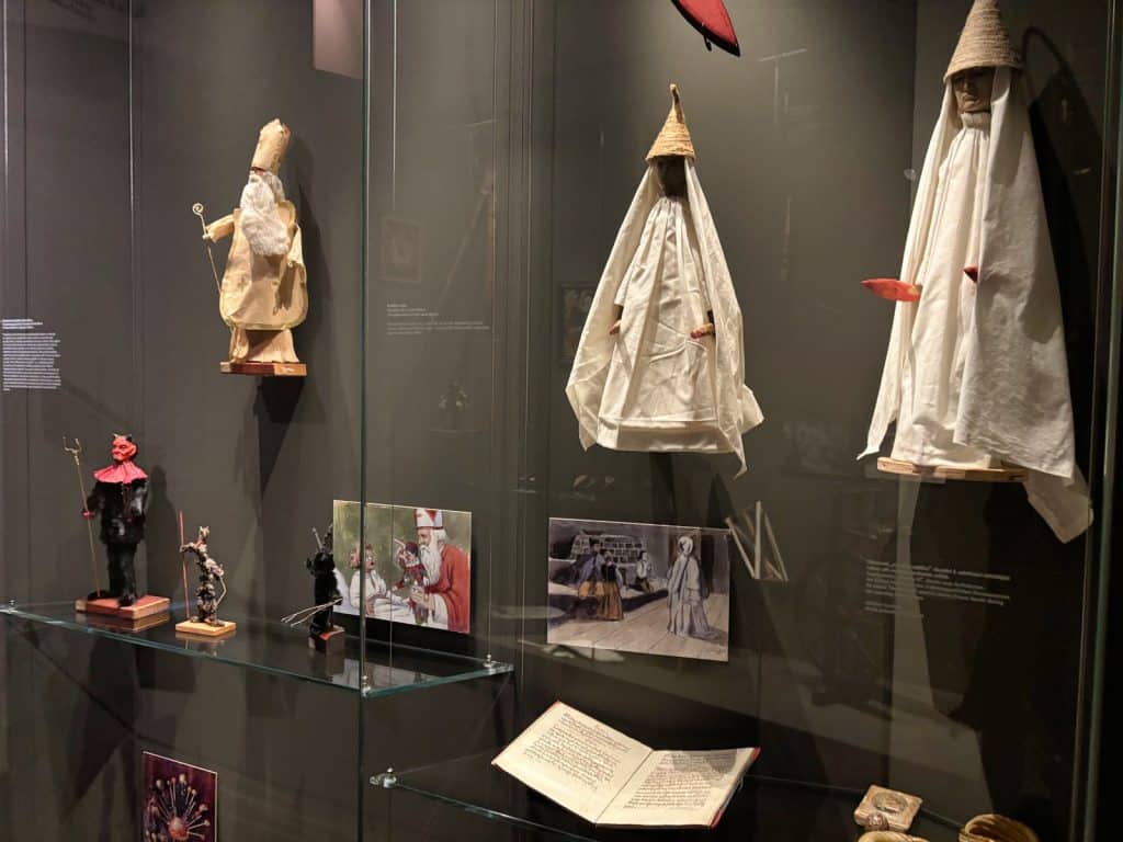 Eine Museumsvitrine im Ethnografischen Museum der Region Pilsen zeigt traditionelle tschechische Weihnachtsfiguren wie den Heiligen Nikolaus, Teufel und weiß gekleidete Gestalten sowie historische Illustrationen und ein aufgeschlagenes Buch.