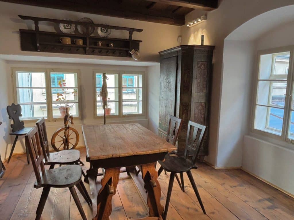 Eine rustikale Wohnstube im Ethnografischen Museum der Region Pilsen mit einem massiven Holztisch, traditionellen Holzstühlen, einem historischen Schrank und einem Spinnrad vor hellen Fenstern. Über den Fenstern hängt ein dunkles Holzregal mit verzierten Tellern und Krügen an einer weiß getünchten Wand unter einer dunklen Balkendecke.