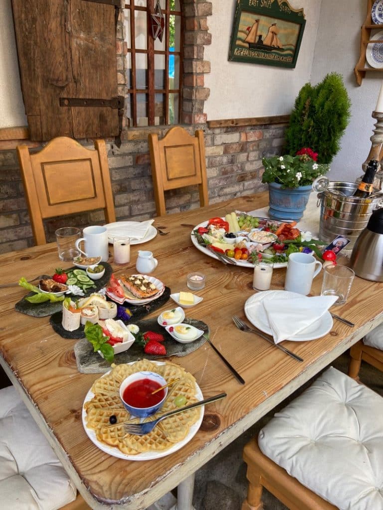 Ein reichhaltig gedeckter Frühstückstisch im Paul's Fantasia Boutique Suites in Stettin bietet eine Auswahl an Waffeln mit Erdbeersauce, Lachs-Wraps, gefüllten Eiern, frischem Obst und einer großen Platte mit Käse und Aufschnitt in rustikalem Ambiente.