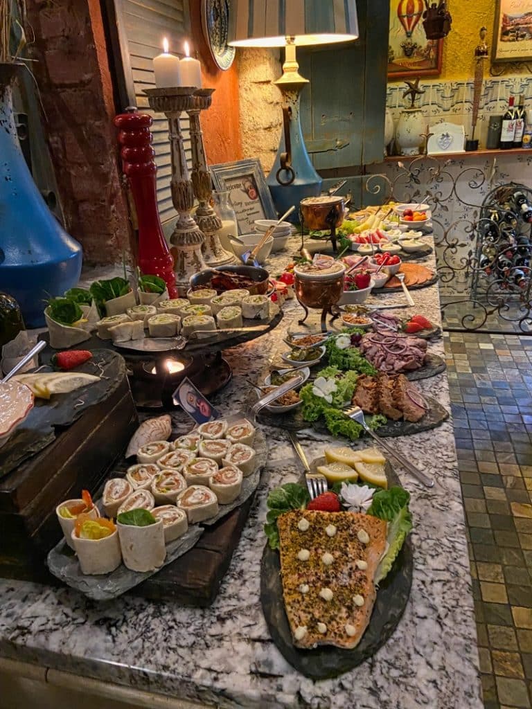 Ein reichhaltiges Frühstücksbuffet im Hotel Paul's Fantasia Boutique Suites in Stettin präsentiert verschiedene Vorspeisen wie Lachsrollen, Wraps, gebratenen Fisch mit Kräuterkruste und Aufschnittplatten auf einer Marmortheke. Im Hintergrund ergänzen rustikale Dekorationen, brennende Kerzen und eine Auswahl an Obst und Salaten das gemütliche Ambiente.