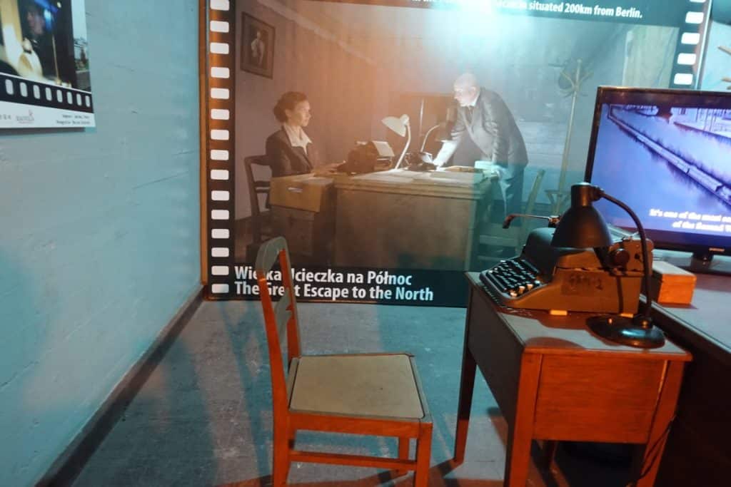 Eine nachgestellte Filmszene im Stettiner Bunker unter dem Hauptbahnhof zeigt einen Schreibtisch mit Schreibmaschine und Lampe vor einem großen Filmplakat mit der Aufschrift „The Great Escape to the North“. Ein hölzerner Stuhl steht im Vordergrund des spärlich beleuchteten Raums, während rechts ein Monitor eine Szene abspielt.