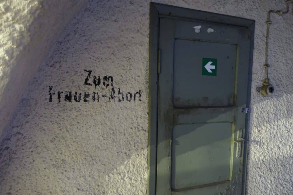 Ein grauer Metalleingang in einem Bunker unter dem Stettiner Hauptbahnhof mit der schwarzen Aufschrift „Zum Frauen-Abort“ an der grob verputzten, weißen Wand.