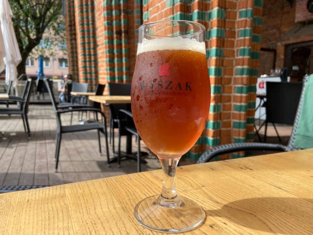 Ein Glas bernsteinfarbenes Wyszak Amber Bier steht auf einem Holztisch im Außenbereich der Brauerei in Stettin vor einer markanten Backsteinmauer.