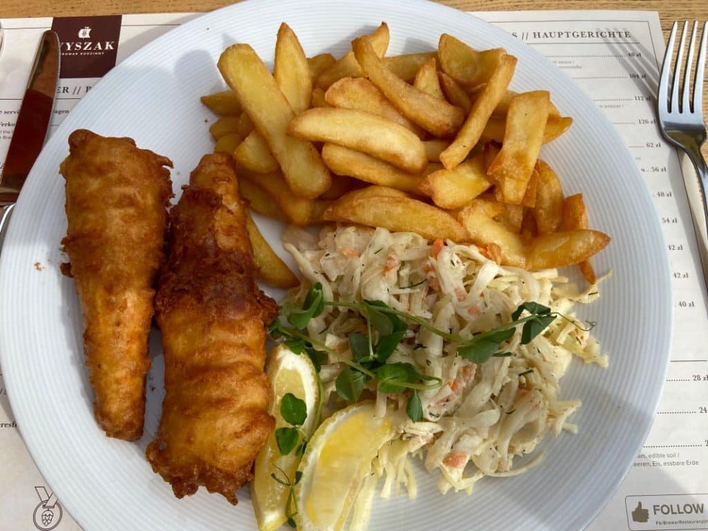 Ein weißer Teller mit Fish and Chips aus der Stettiner Brauerei Wyszak, bestehend aus zwei goldbraun frittierten Fischfilets, einer Portion Pommes frites, Krautsalat und Zitronenspalten.
