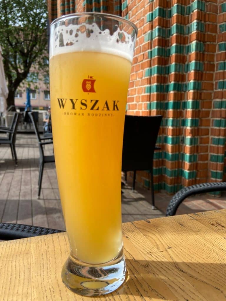 Ein hohes Glas naturtrübes Wyszak Weizenbier steht auf einem Holztisch im Außenbereich der Wyszak Brauerei in Stettin vor einer markanten rot-grünen Backsteinwand.