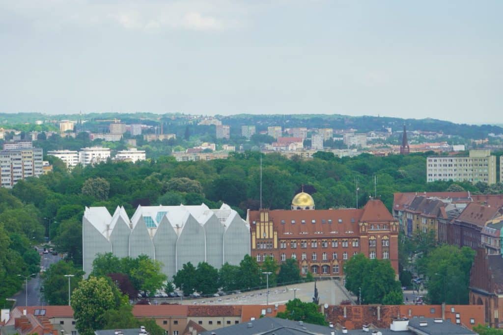 Blick von einem Aussichtspunkt in Stettin auf die markante, weiße Architektur der Philharmonie und das benachbarte, backsteinerne Polizeipräsidium mit seiner goldenen Kuppel inmitten einer grünen Stadtlandschaft.