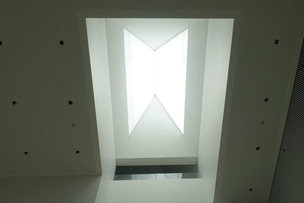 Blick von unten auf ein geometrisch geformtes, helles Dachfenster in der Philharmonie Stettin, das von einer minimalistischen, weißen Deckenkonstruktion mit kleinen Einbaustrahlern umrahmt wird.