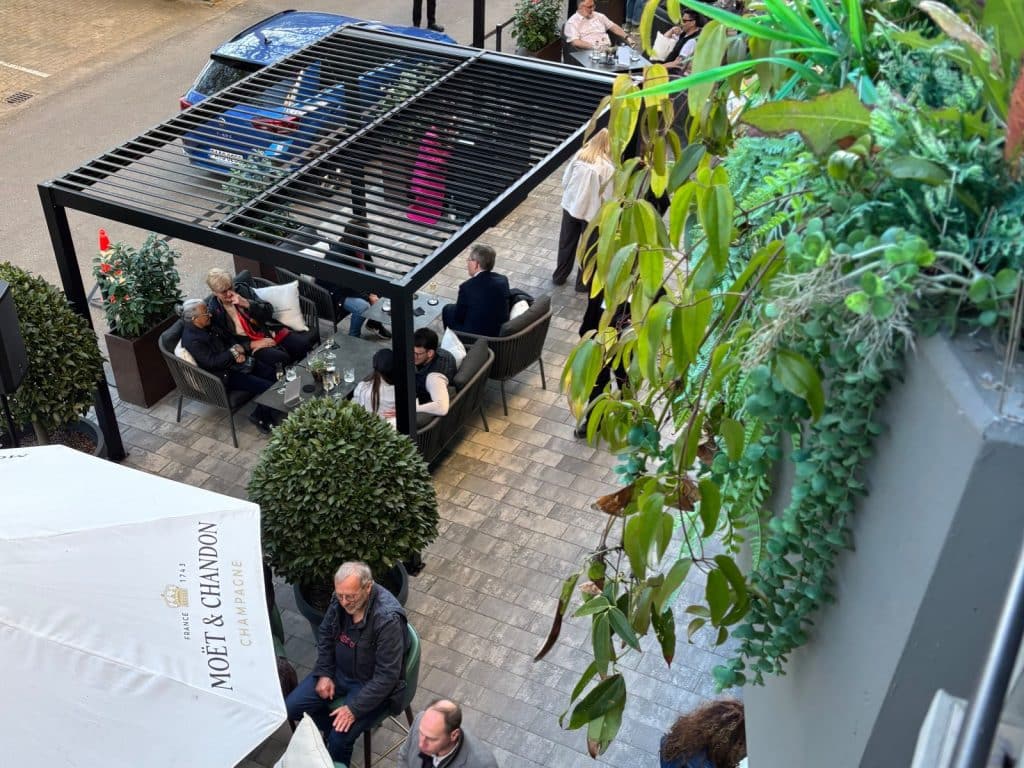 Blick von oben auf die Außenterrasse des DoubleTree by Hilton Berlin Ku'damm, auf der Gäste unter einer schwarzen Pergola und Sonnenschirmen an Tischen sitzen, umgeben von üppigem Grün.